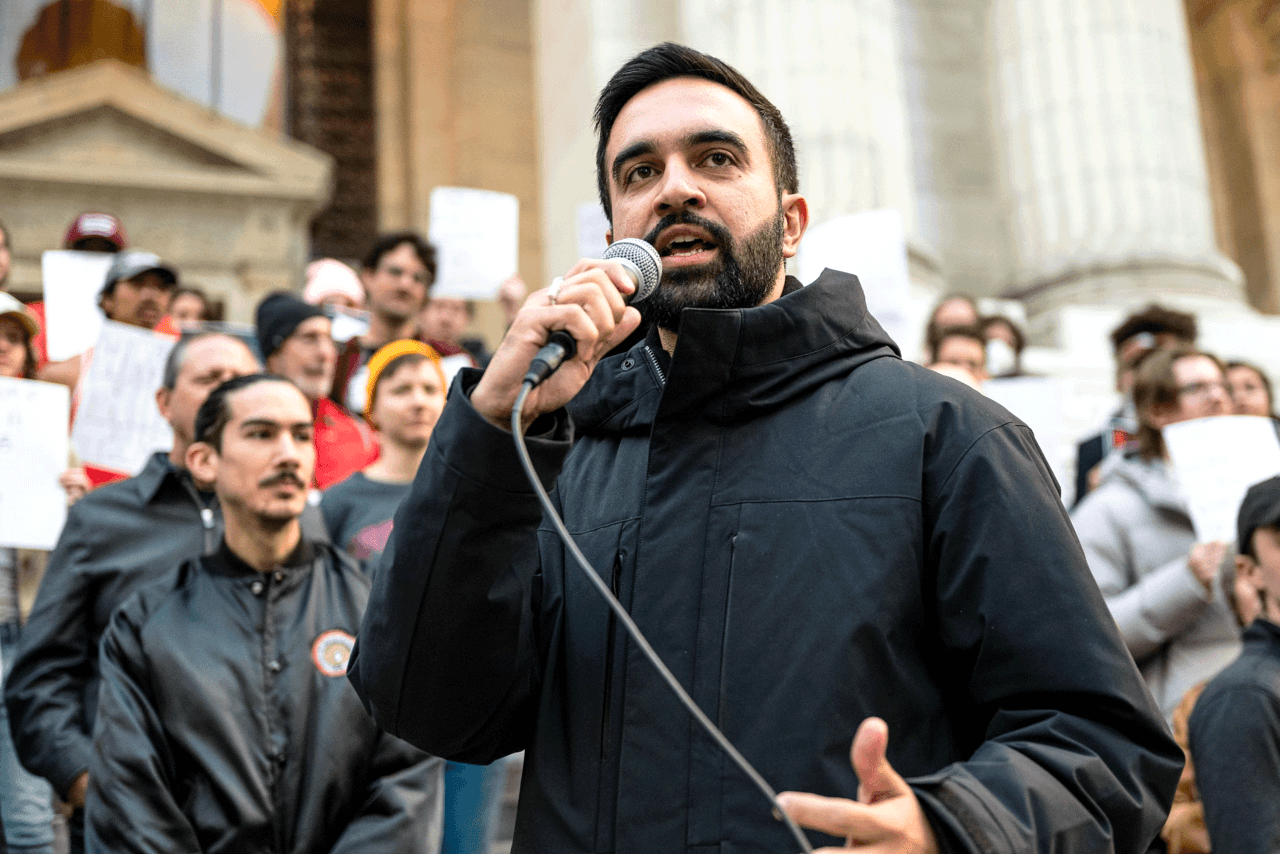 Zohran Mamdani holding a microphone and speaking during a political event