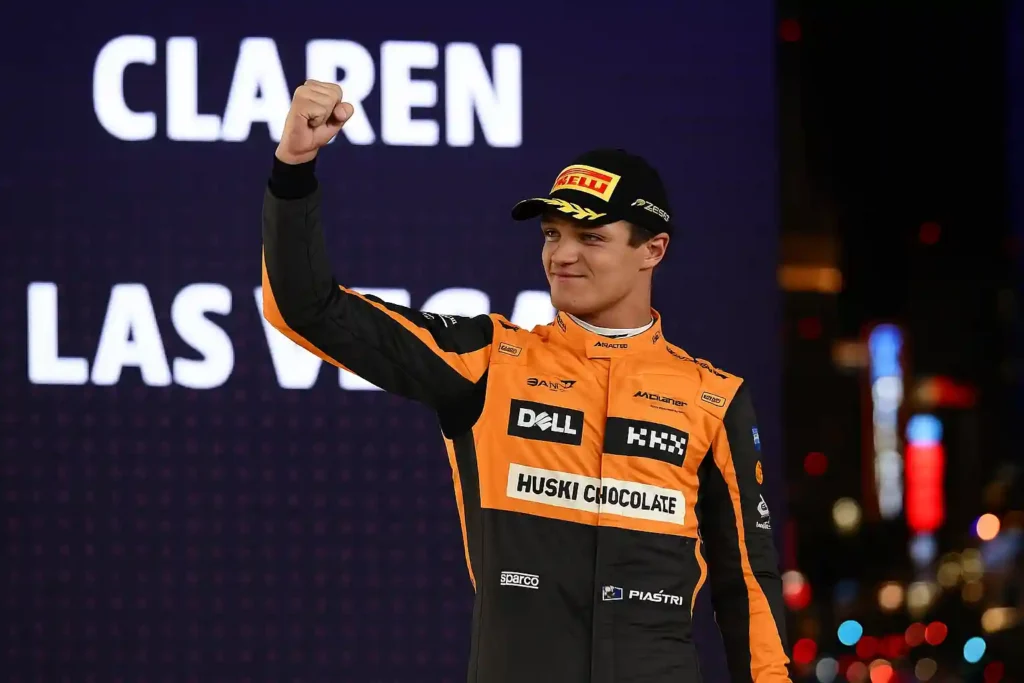 Lando Norris celebrates on the podium under bright neon lights after the Las Vegas Grand Prix, wearing his McLaren race suit and raising his fist in triumph.