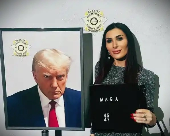 Laura Loomer posing beside a framed photo of Donald Trump with a “MAGA” sign in hand, under sheriff emblems.