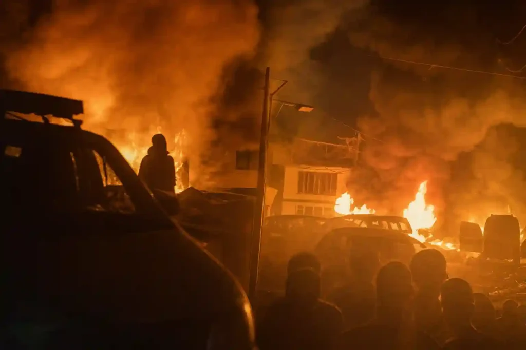Fire engulfs vehicles and a building at night as thick smoke rises, with silhouettes of people watching the blaze from a distance. Nowgam Srinagar blast