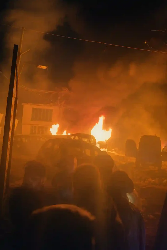 Large nighttime fire engulfs vehicles and a damaged building as thick smoke rises, while silhouettes of people gather at the scene.