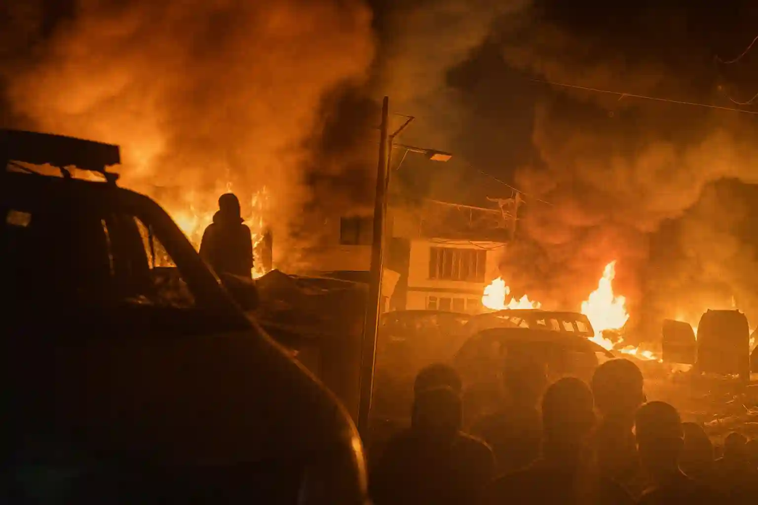 Fire engulfs vehicles and a building at night as thick smoke rises, with silhouettes of people watching the blaze from a distance. Nowgam Srinagar blast