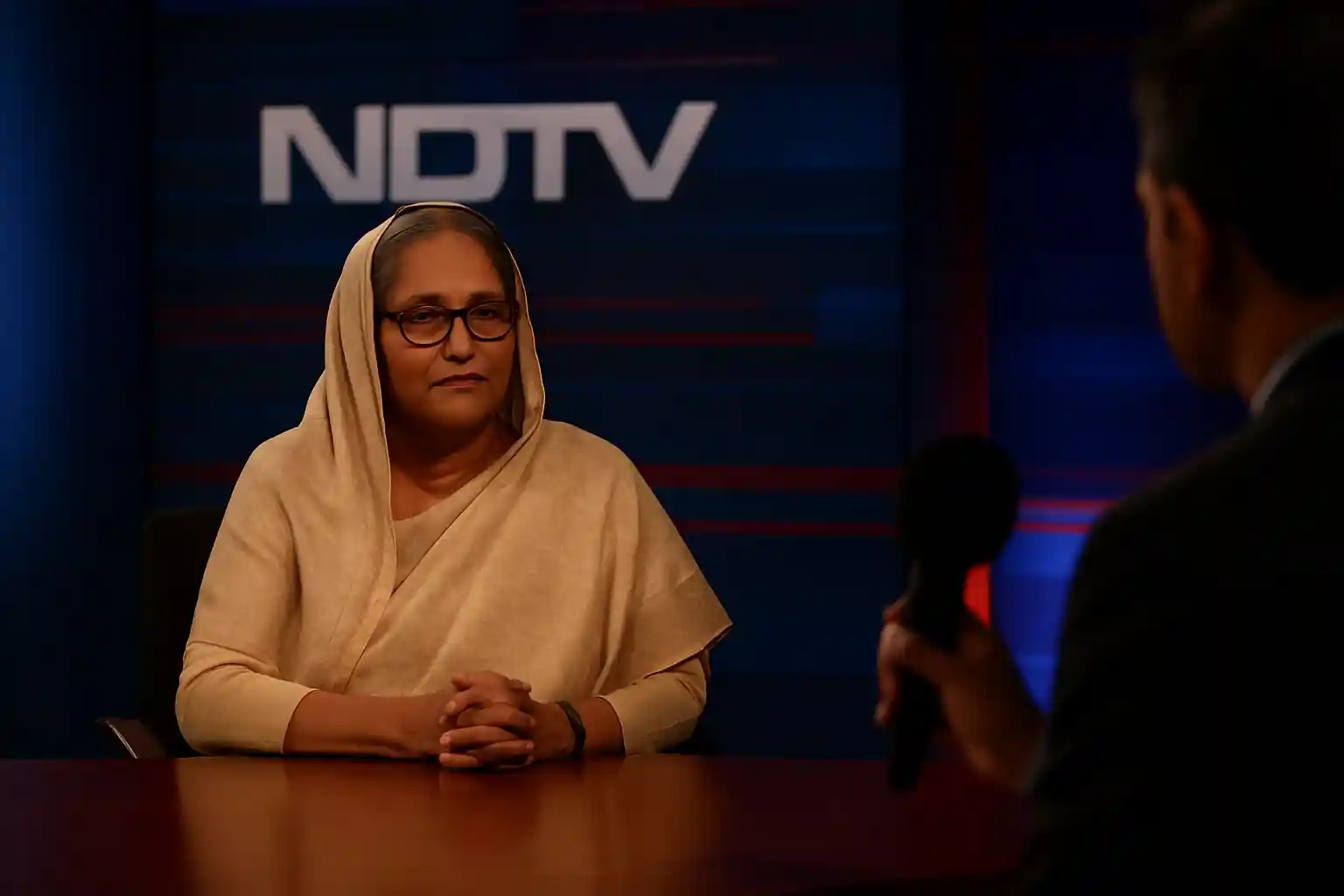 A woman in a saree sits for an NDTV-style discussion scene representing Sheikh Hasina latest interview, captured with soft studio lighting.