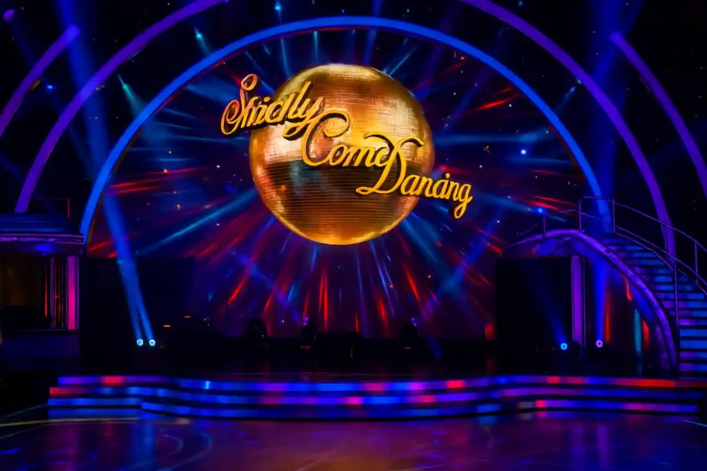 A vibrant Strictly Come Dancing stage with colorful lighting and a golden disco ball, illustrating the show’s setting during coverage of the Strictly rape accusation.