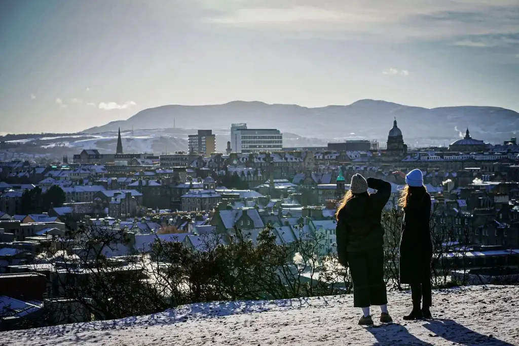 Two people standing on a snowy hill overlooking a winter cityscape, illustrating conditions linked to the UK snow forecast.