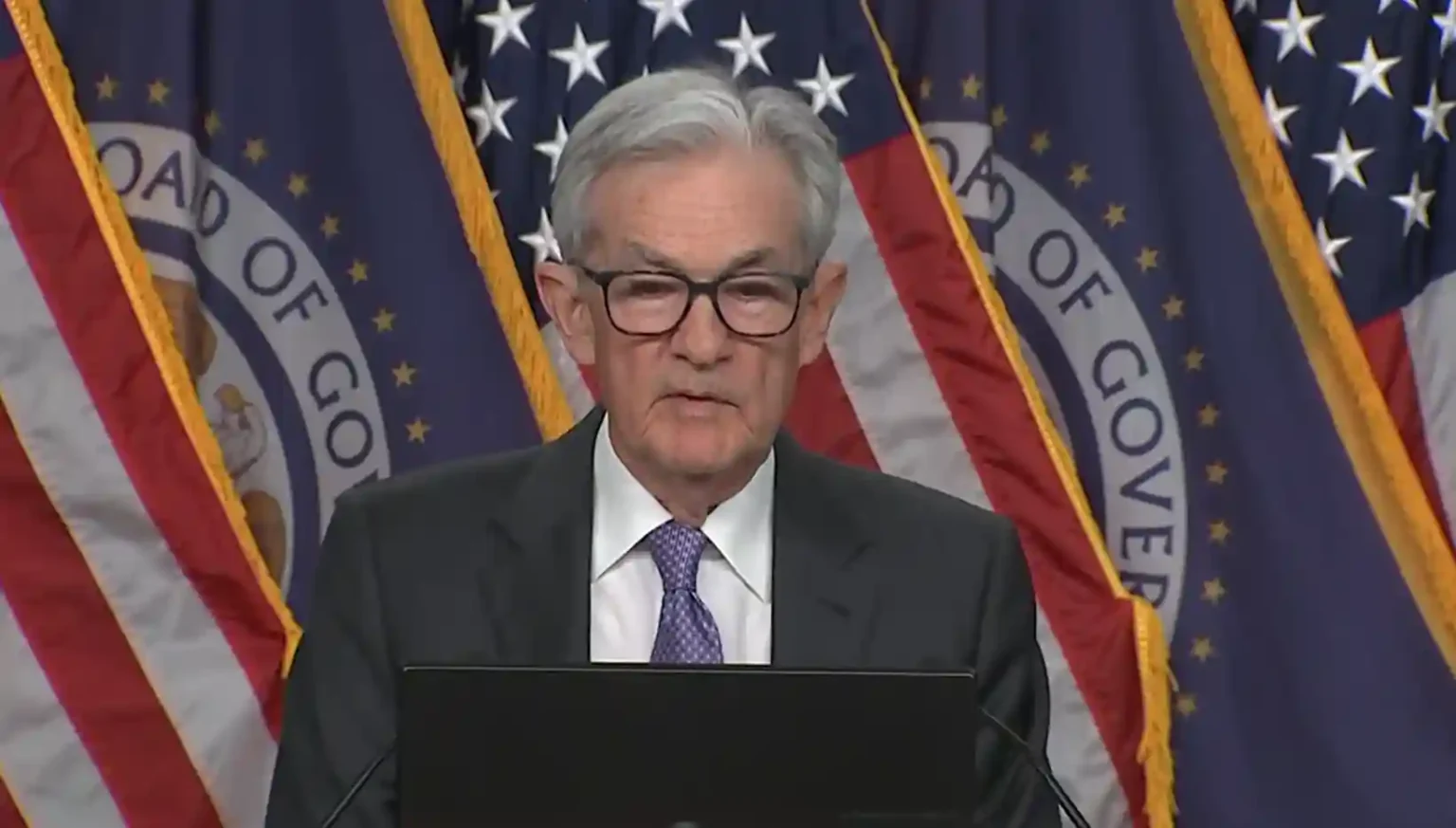 Federal Reserve chair speaking at a press briefing with U.S. flags in the background during an announcement on the Powell interest rate cut.