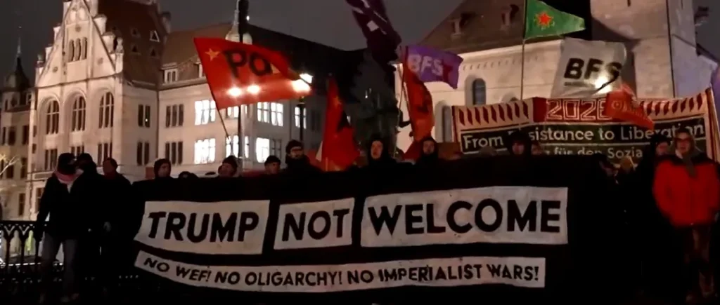 Protesters hold a banner reading “Trump Not Welcome” during a demonstration against the World Economic Forum, with flags and historic buildings visible in the background.